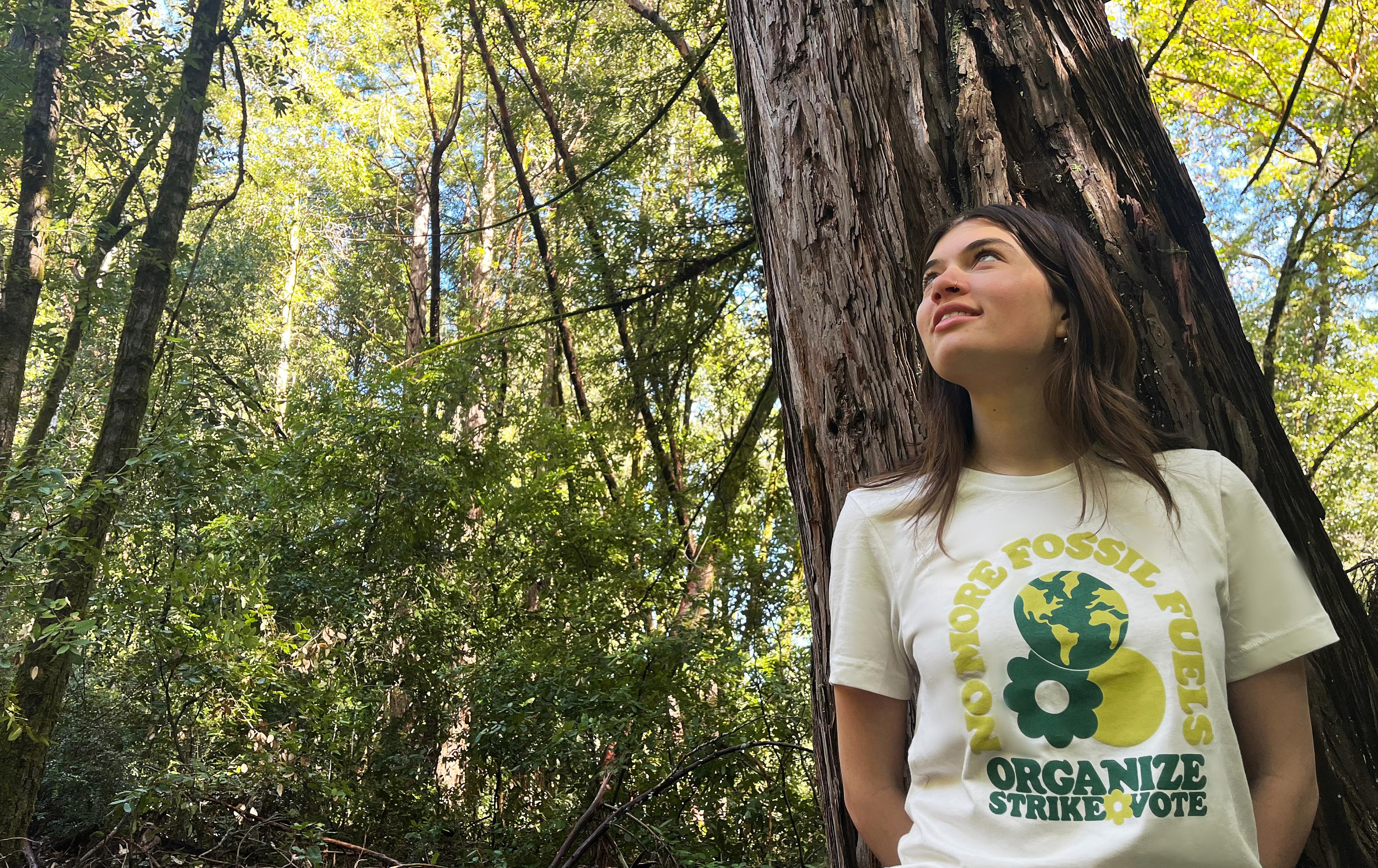 
        Model standing in front of a tall tree.
        She is looking up into the leaves and wearing the organize tee.
      